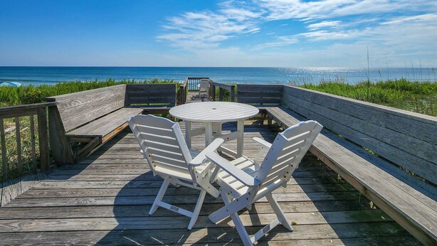 Une terrasse en bois avec une table ronde et quatre chaises blanches, donnant sur une plage.