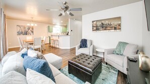 Een moderne woonkamer met een bank, een poef, en fauteuils, een eethoek met een tafel en stoelen, en een keuken op de achtergrond.
