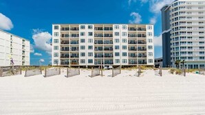 Um hotel à beira-mar com vários andares e varandas, uma praia de areia em primeiro plano, e um céu azul e limpo.