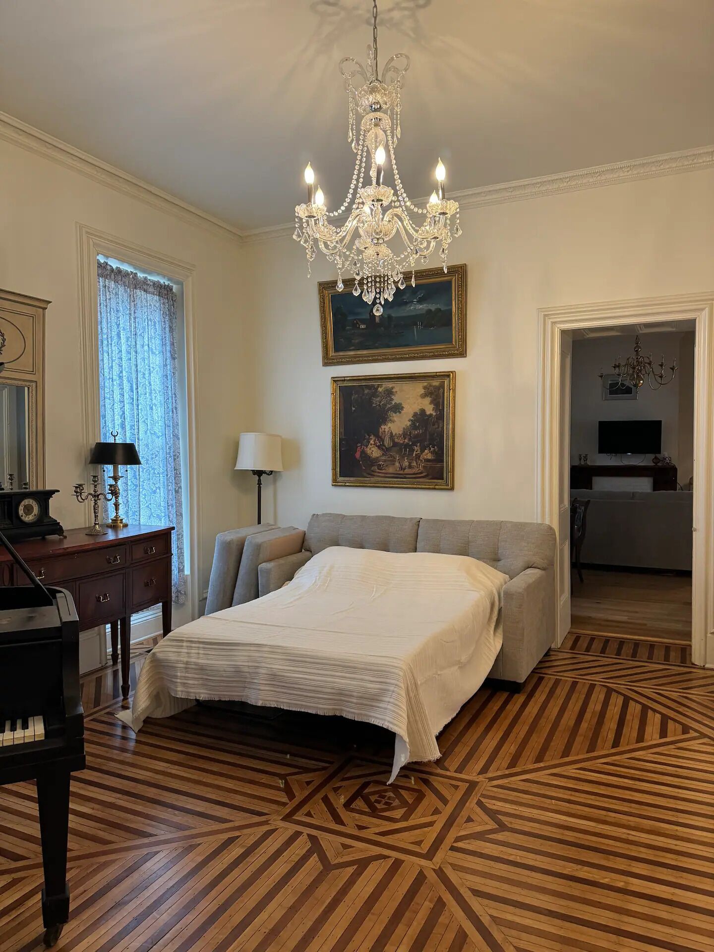 A room with a bed, a chandelier, a piano, and a painting on the wall.