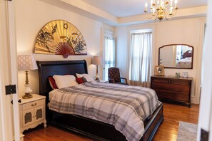 Une chambre à coucher comprenant un lit, des tables de chevet, une commode, une chaise, un lustre et une grande œuvre d’art murale.