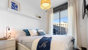 A modern bedroom with a large bed, a bedside table with a lamp, a wall-mounted shelf with decorative items, and a view of buildings through the window.