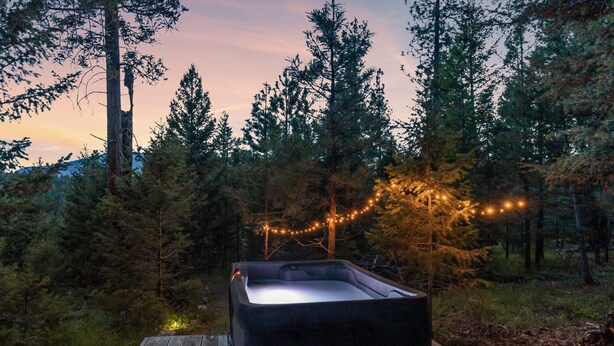 Un bain à remous sur une terrasse en bois, entouré d’arbres, avec des guirlandes lumineuses en arrière-plan.
