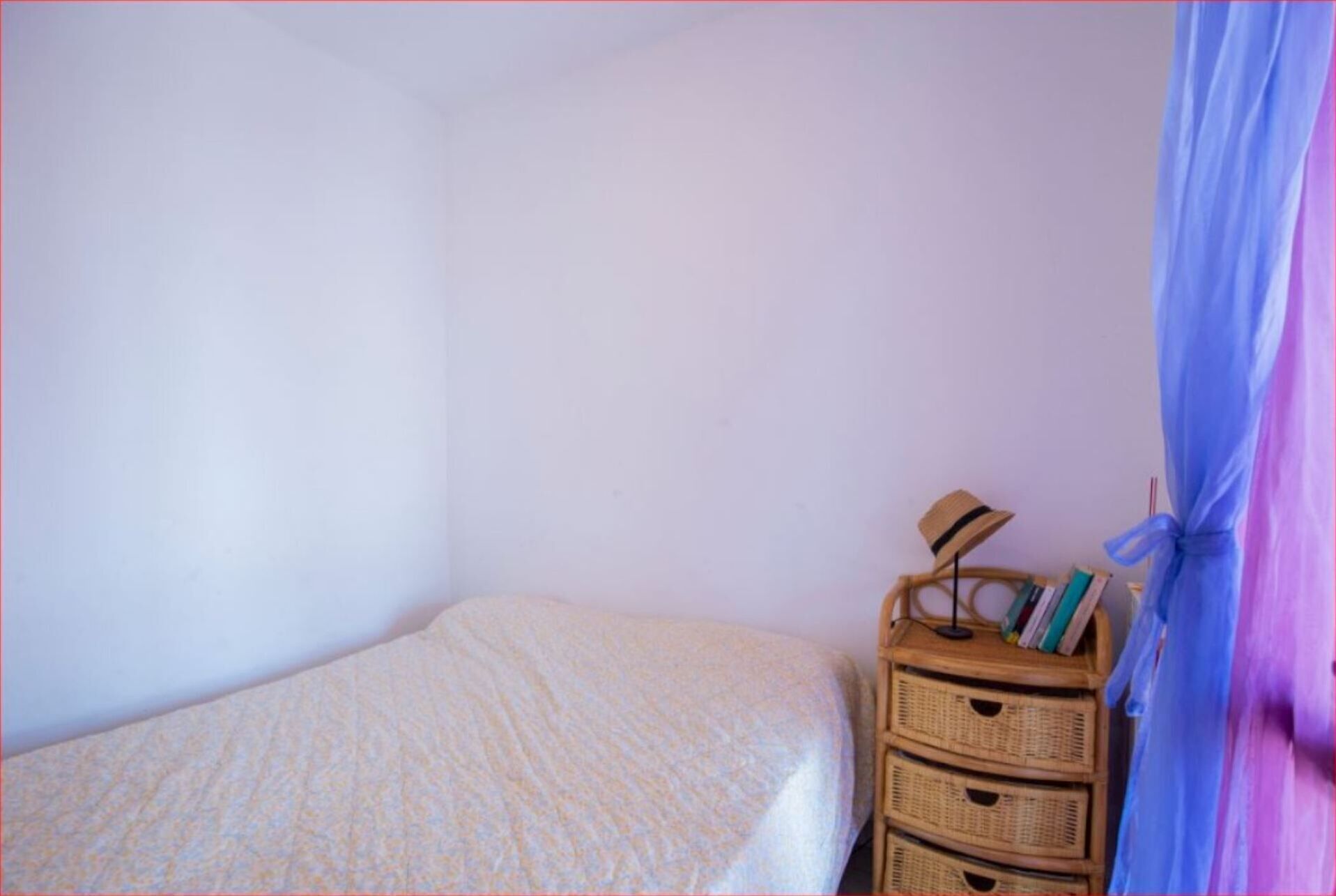 A bedroom with a single bed, a wooden dresser with books and a hat, and a blue curtain.