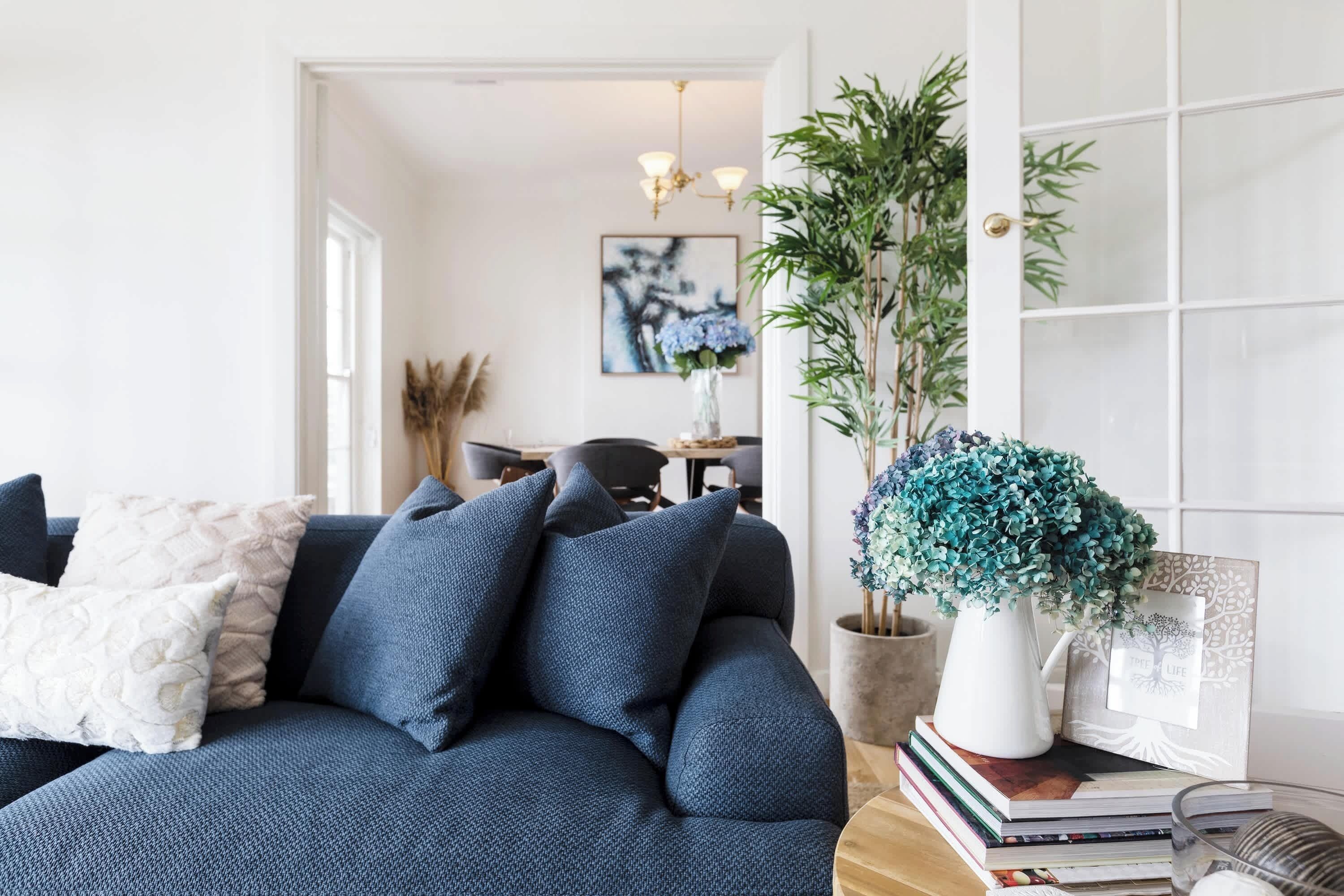 Een woonkamer met een donkerblauwe bank, een witte vaas met blauwe bloemen, en een ingelijst beeld op een tafel.