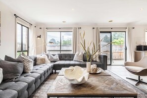 A modern living room with a sectional sofa, a coffee table, and a view through large windows. - Provincial 4BR Family Retreat Near Train & Shops (South Morang)