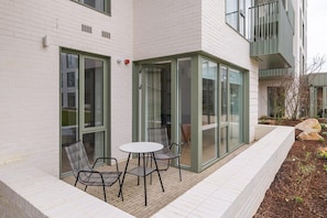 A modern building with a glass door entrance, a small patio with a table and chairs, and a brick pathway.