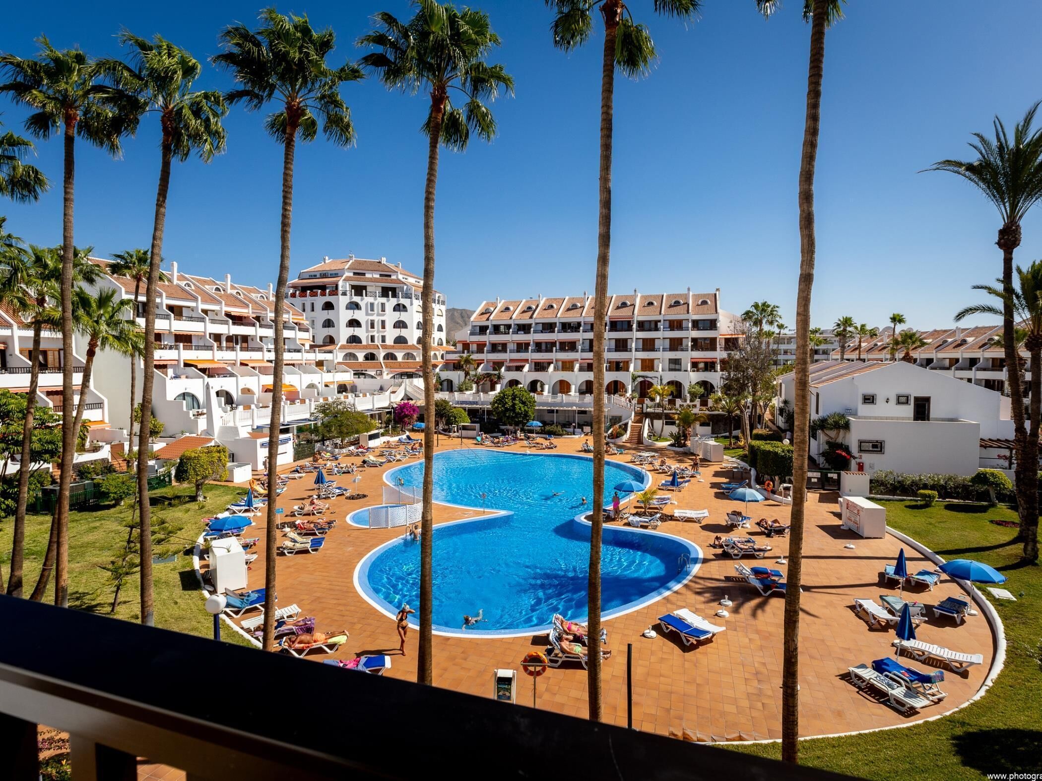 A resort with a large central pool, multiple guest buildings, and palm trees.