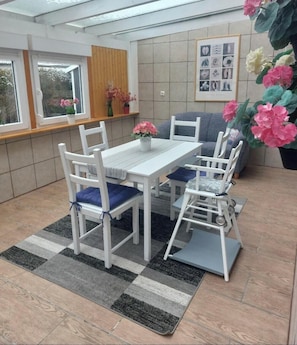 A dining area with a table and chairs, a rug, and a window with flowers.