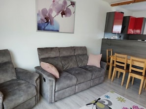 A living room with a grey sofa, a wooden dining table with chairs, a kitchen area with red cabinets, and a framed picture on the wall.
