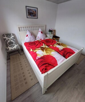 A bedroom with a white bed, a patterned chair, a nightstand with a plant, and a framed picture on the wall.