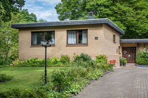 A two-story brick house with a paved driveway, a small garden with various plants, and a mailbox.