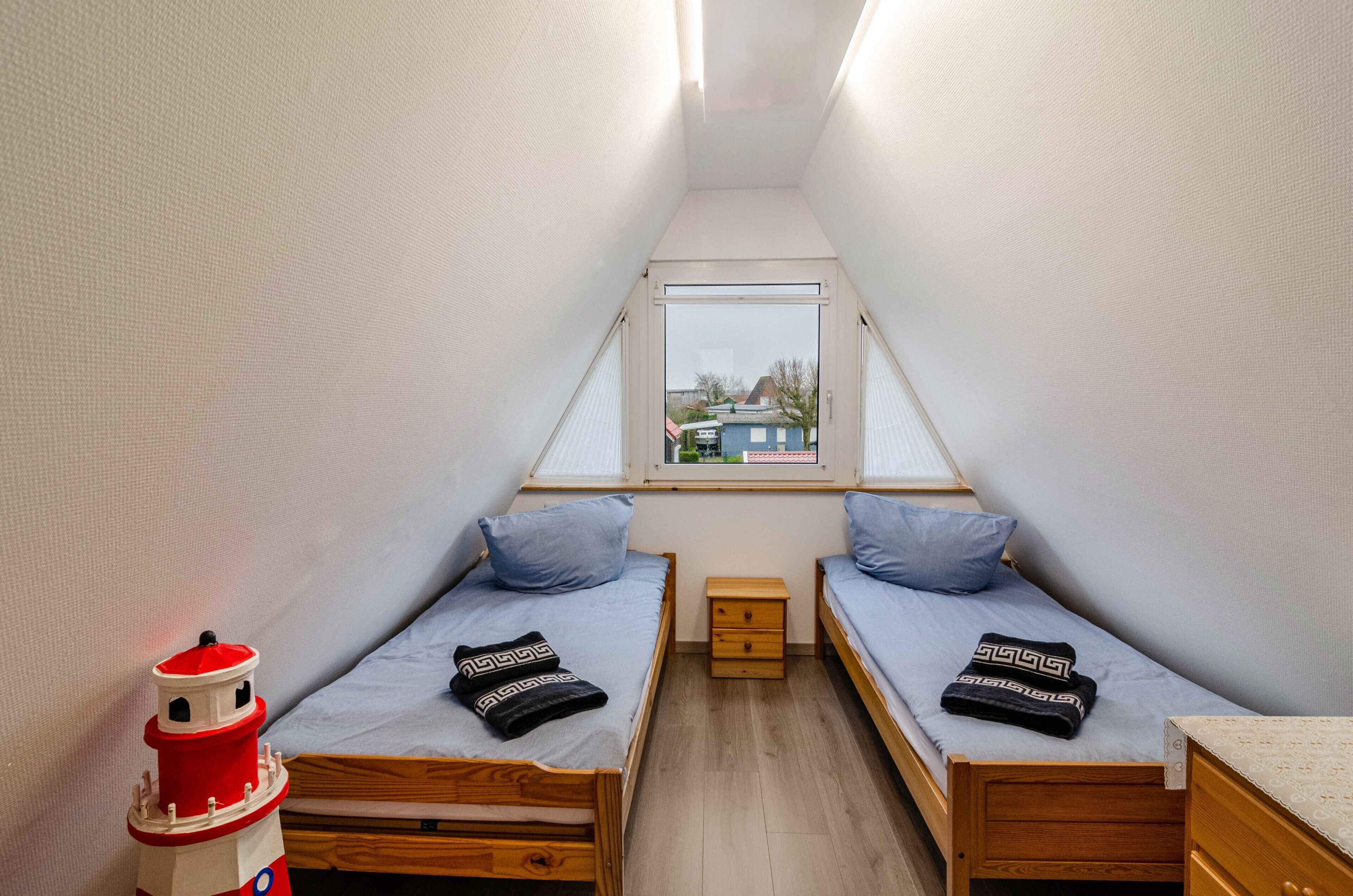 Una habitación pequeña y estrecha con dos camas individuales, una mesita de noche de madera, un ventanal pequeño y un adorno de faro rojo y blanco.