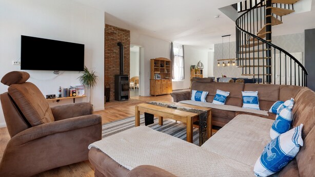 Un salon moderne avec un canapé, un fauteuil et une table basse en bois. Un téléviseur est fixé au mur, et on peut voir un escalier avec une rampe en bois.