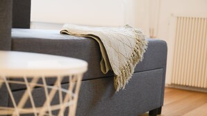 Ein graues Sofa mit einer beigen Tagesdecke, ein runder weißer Tisch mit Geflechtuntergestell und ein Holzboden.