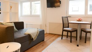 Ein modernes Wohnzimmer mit einer Couch, einem Esstisch mit Stühlen, einem an der Wand montierten Fernseher und einem Fenster mit Vorhängen.