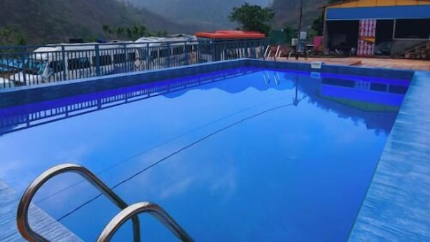 A swimming pool with a metal ladder, surrounded by a fence, with mountains in the background. - Rudra Adventures (Narendranagar)