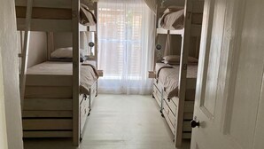 Quarto pequeno com duas beliches, uma porta e uma janela com cortinas leves.