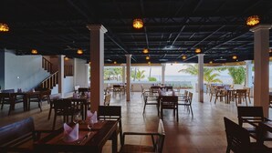 A restaurant with a terrace overlooking the sea, featuring wooden tables and chairs, and a view of palm trees and the ocean.