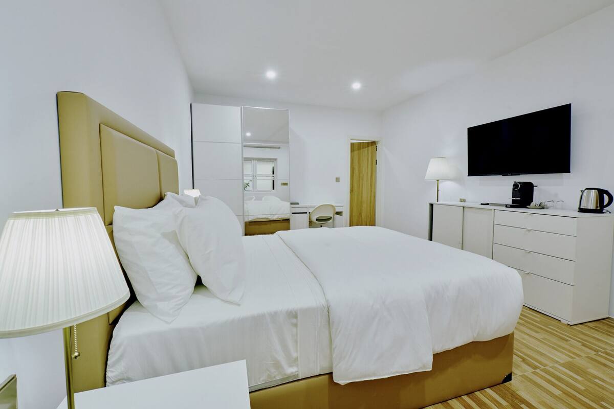 Quarto de hotel moderno com uma cama grande, mesas de cabeceira, escrivaninha, cadeira, televisão e máquina de café.