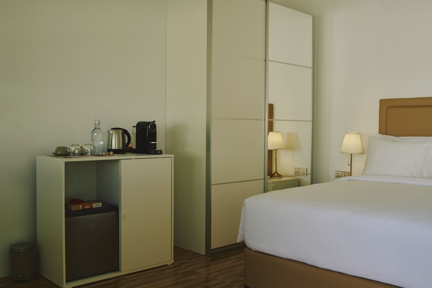 Een hotelkamer met een bed, nachtkastje met lamp, minibar en een spiegel.