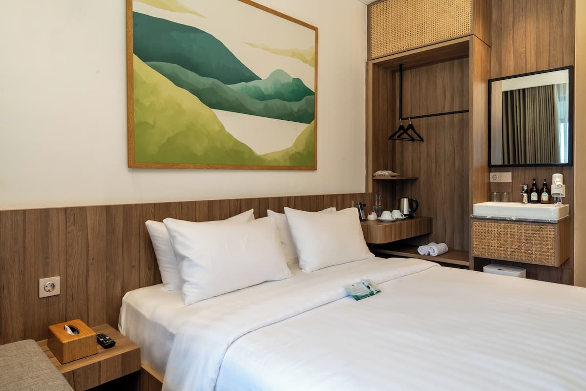 Habitación de hotel con cama, mesita de noche, un cuadro en la pared, armario de madera, espejo y un pequeño baño con lavamanos y artículos de aseo.