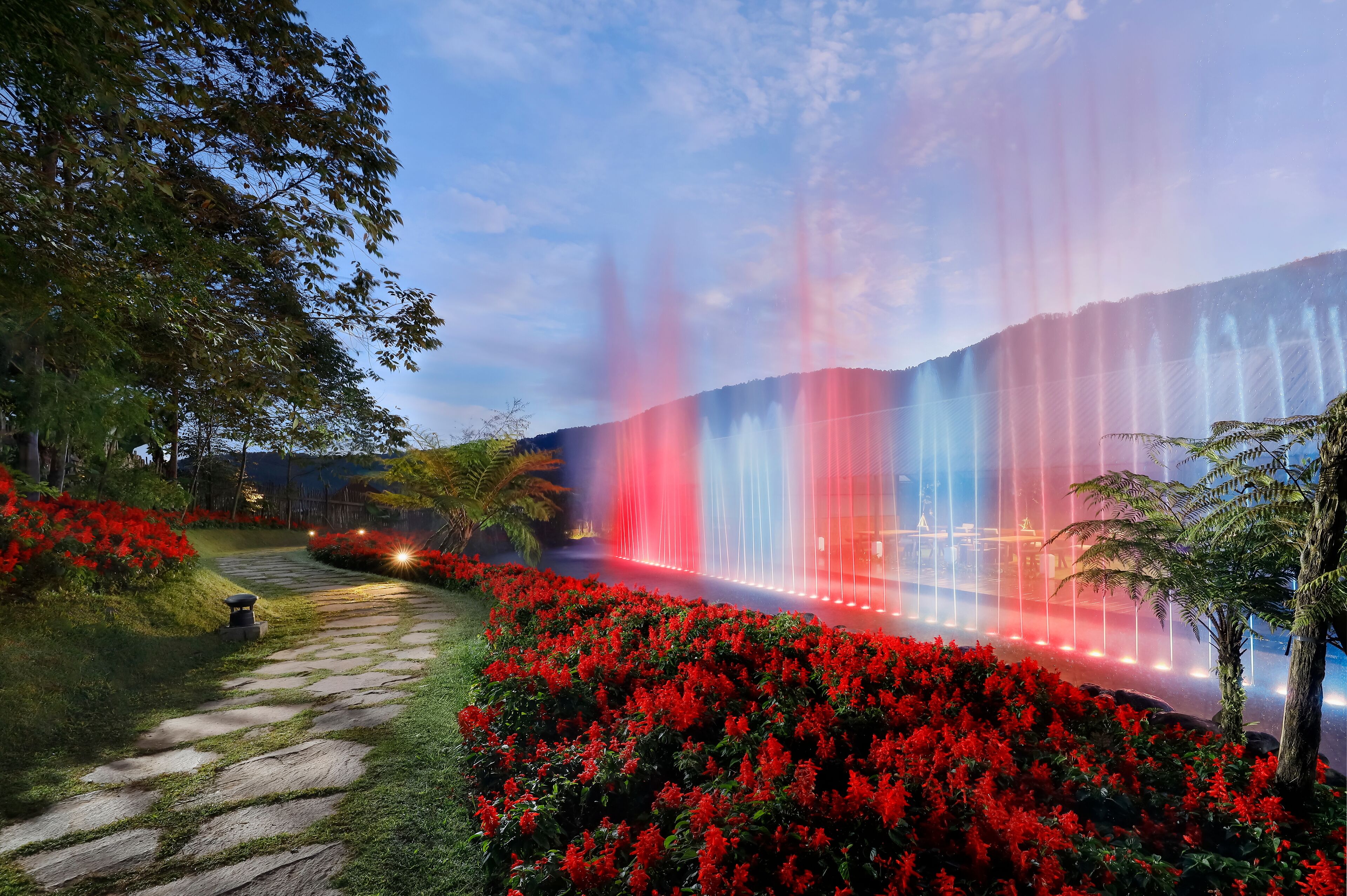 A garden with a stone pathway, red flowers, and a fountain with water jets.