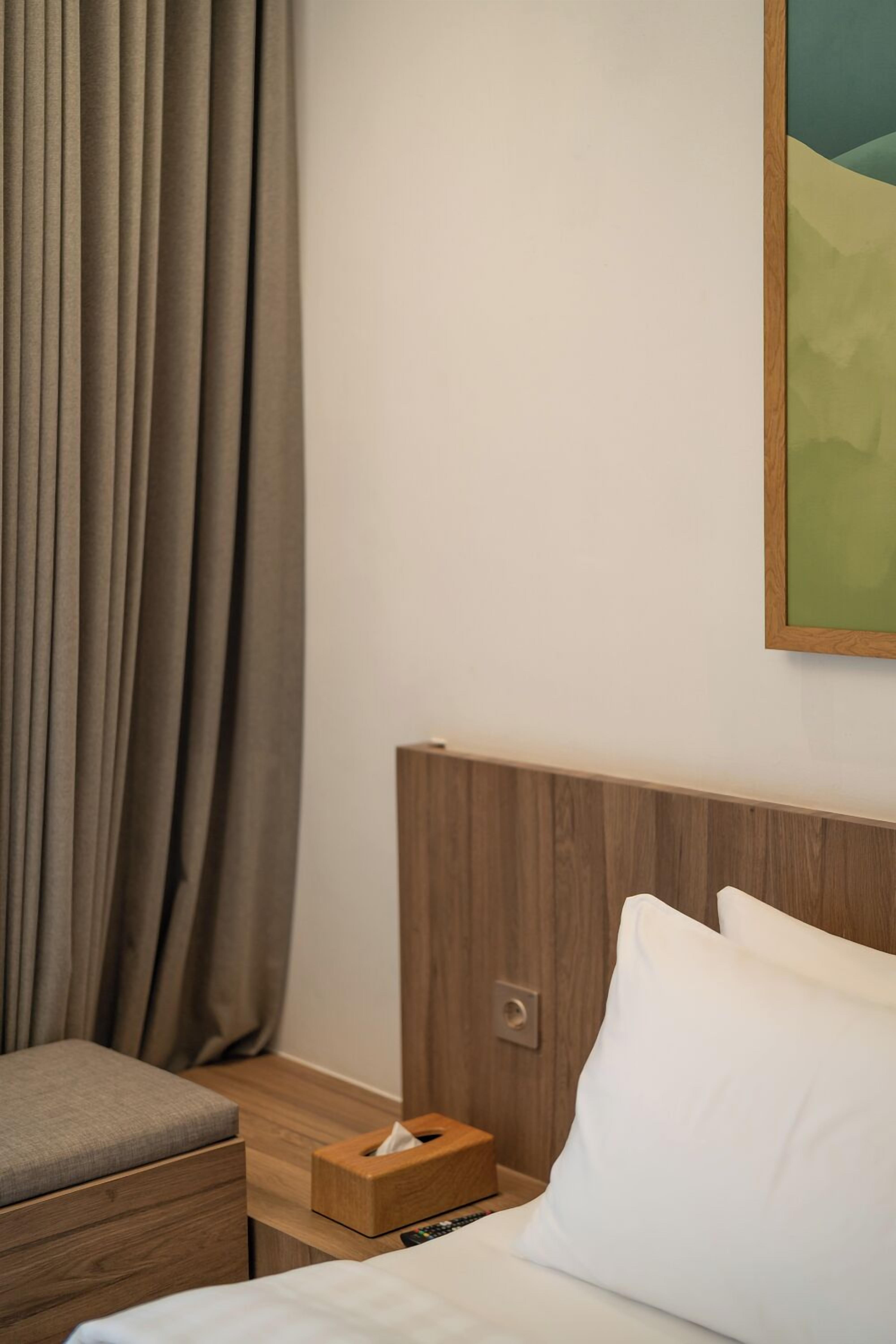 A hotel room with a wooden headboard, a bedside table with a tissue box, a bed with white linens, and a view of a green landscape through the window.