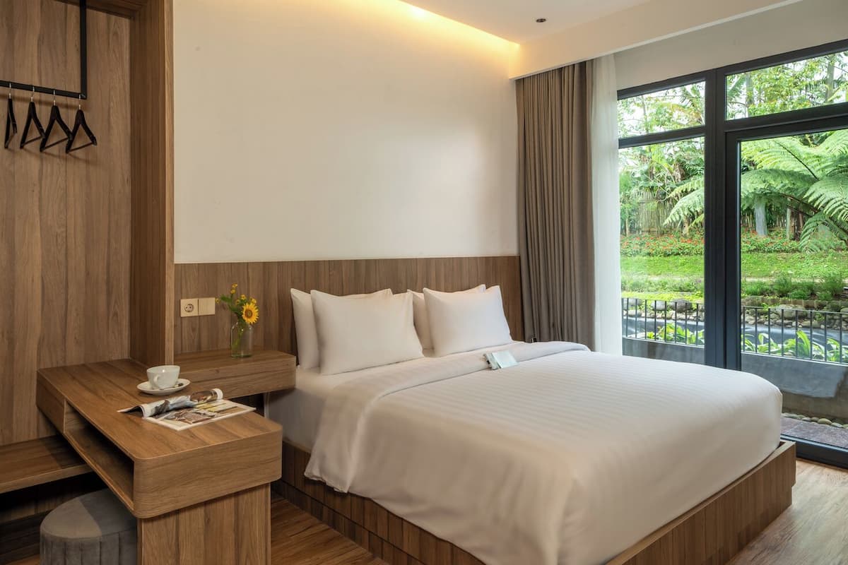 Habitación de hotel moderna con una cama grande, una mesita de noche de madera, una taza de café y vistas a un jardín exuberante a través de amplias ventanas.