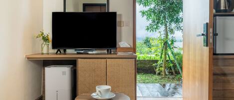 Habitación moderna con televisor de pantalla plana sobre una consola de madera, una pequeña mesa con una taza y vistas a un jardín a través de una puerta abierta.