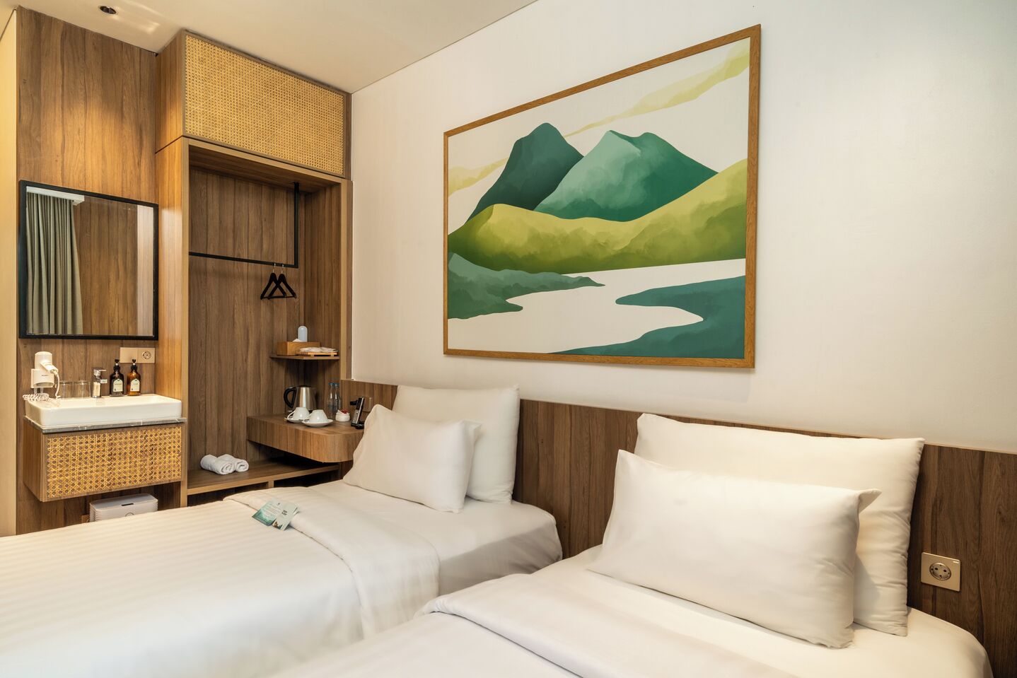 A hotel room with two beds, a wooden wardrobe, a bathroom with a sink and mirror, and a painting of mountains.