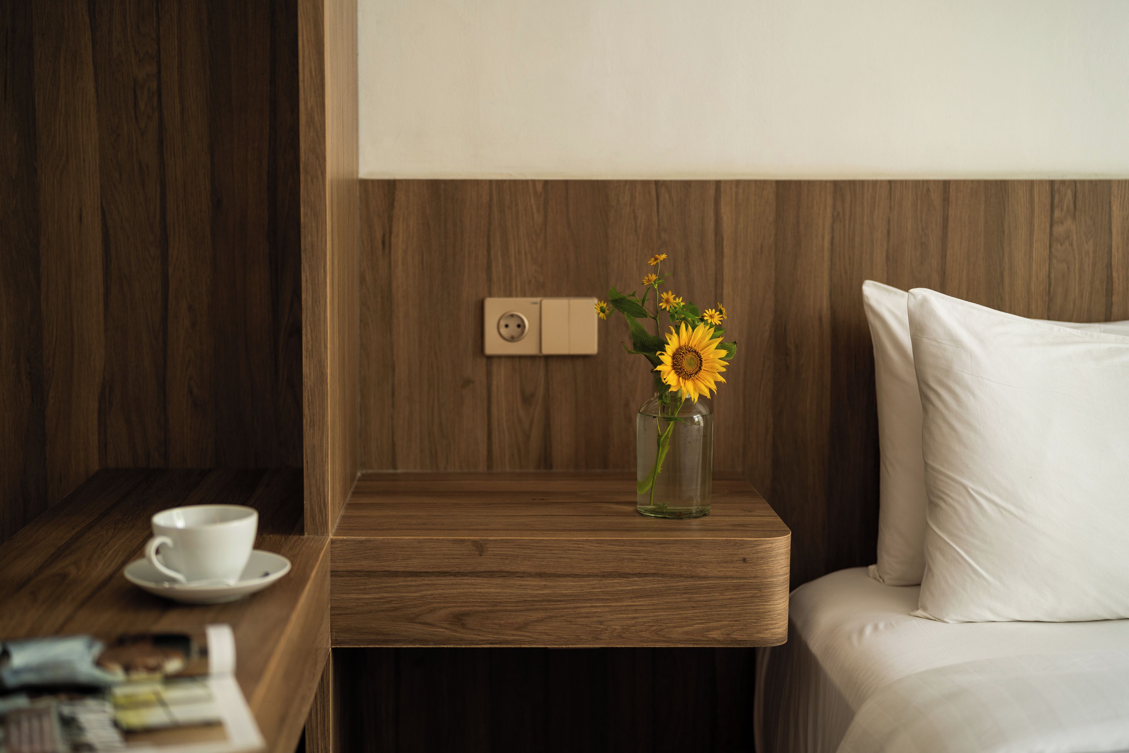 Una mesita de noche de madera con una taza y platillo blancos, un florero con girasoles y un enchufe.