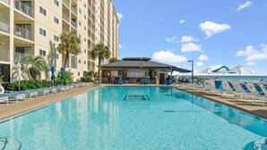 A swimming pool with lounge chairs and umbrellas, a building with balconies, and a beach area with umbrellas.