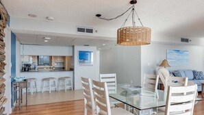Una cocina moderna con una isla central, un comedor con mesa de vidrio y sillas blancas, y una sala de estar con sofá y cojines.