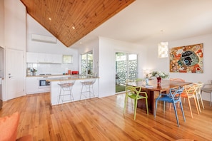 Une cuisine moderne avec des meubles blancs, un plafond en bois et un coin repas agrémenté de chaises colorées et d’une table en bois.