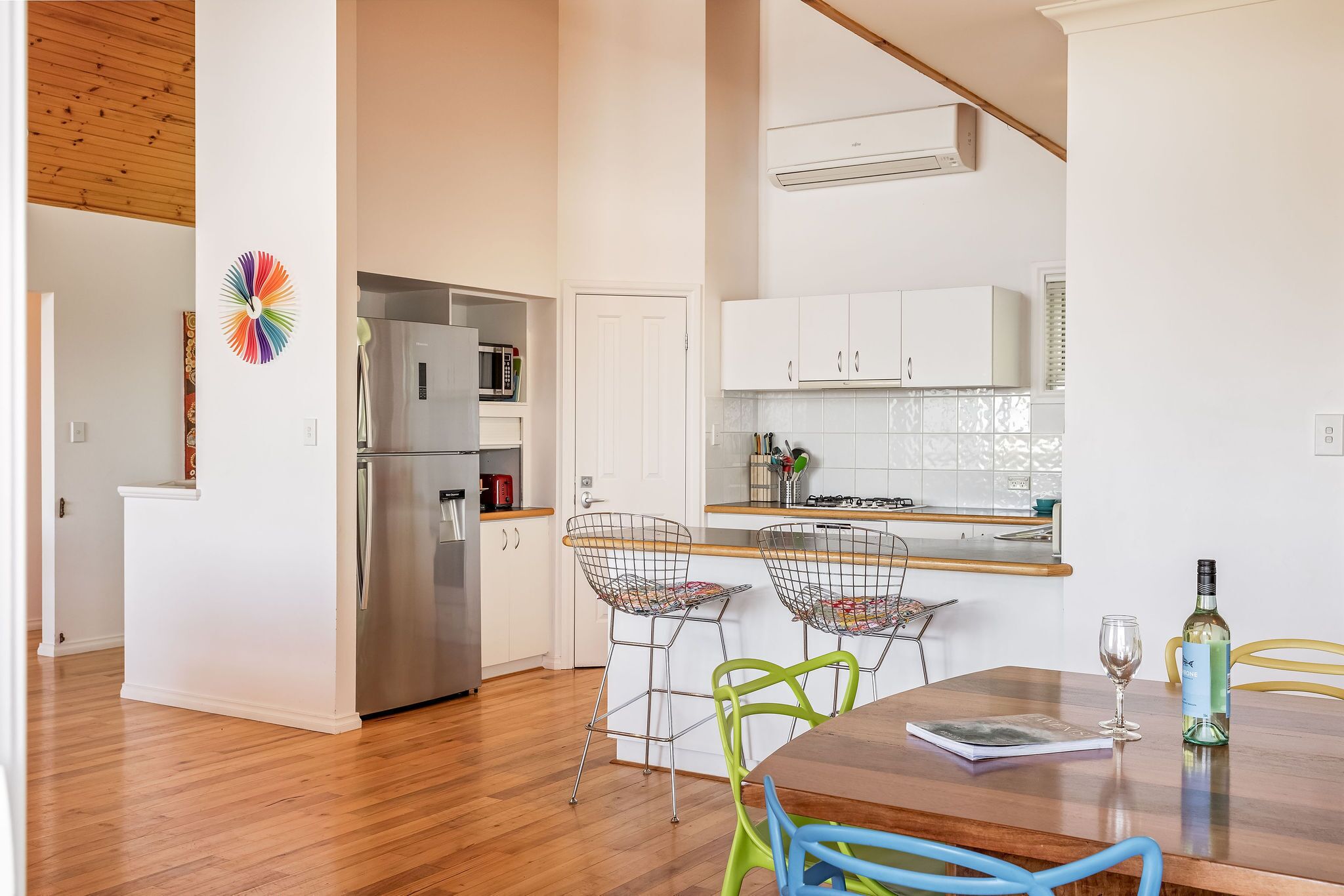 A modern kitchen with white cabinets, a stainless steel refrigerator, and a wooden dining table with colorful chairs.