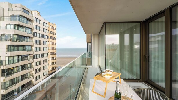 A balcony with a table set for two, overlooking a beach and adjacent buildings. - Sol i Mar - located in the heart of Westende (Middelkerke)