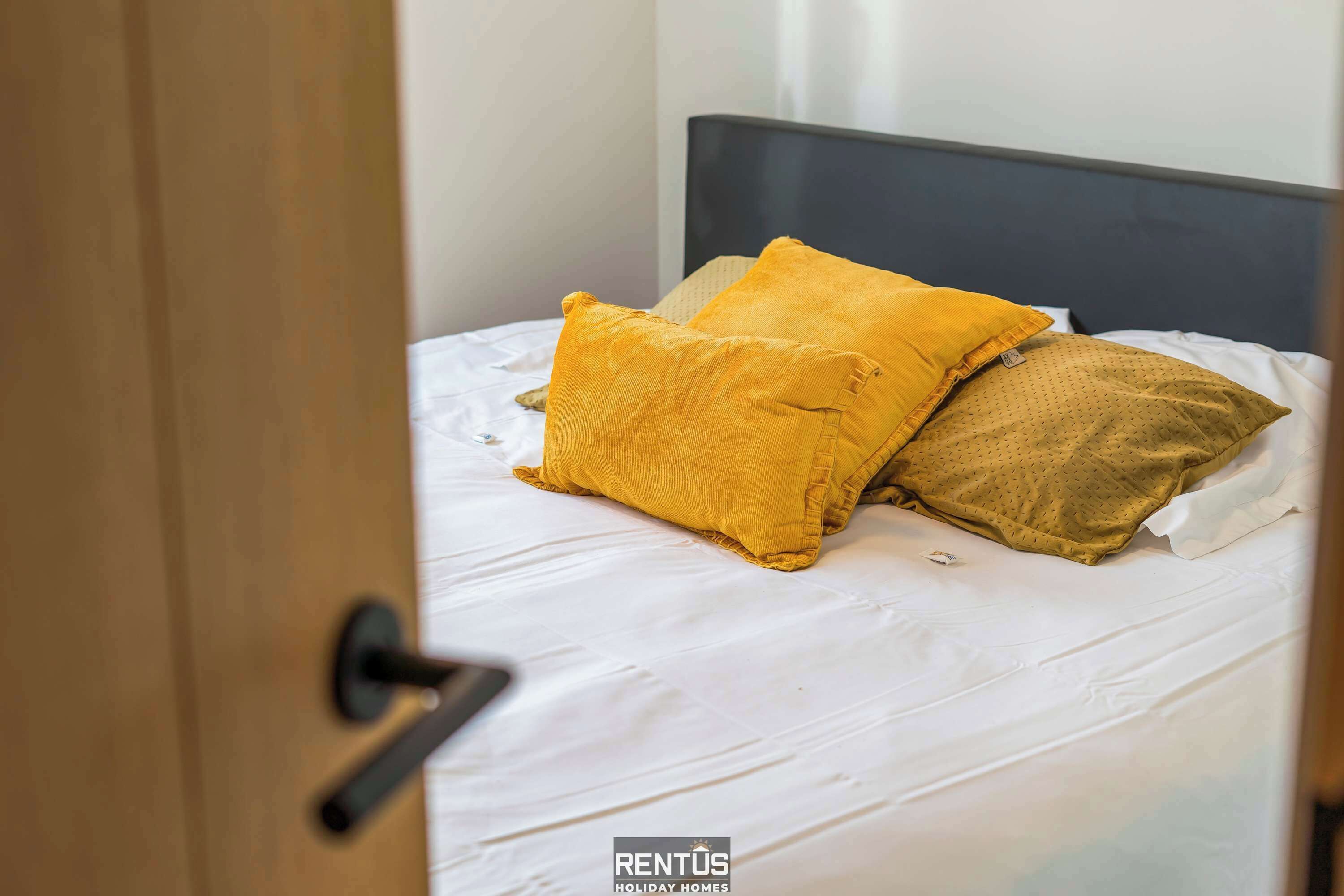 A bed with white bedding and two pillows, one yellow and one brown, in a room with a wooden door and a dark headboard.