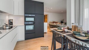 A modern kitchen with white cabinetry, a black refrigerator, and a built-in oven. There is a dining area with a table and chairs, and a window with curtains. - Sol i Mar - located in the heart of Westende (Middelkerke)