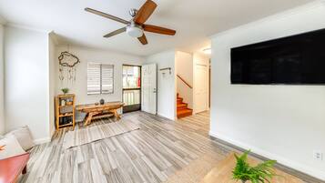 Un salón espacioso con piso de madera, un ventilador de techo, un televisor de pantalla plana, una escalera y una puerta que da a otra habitación.