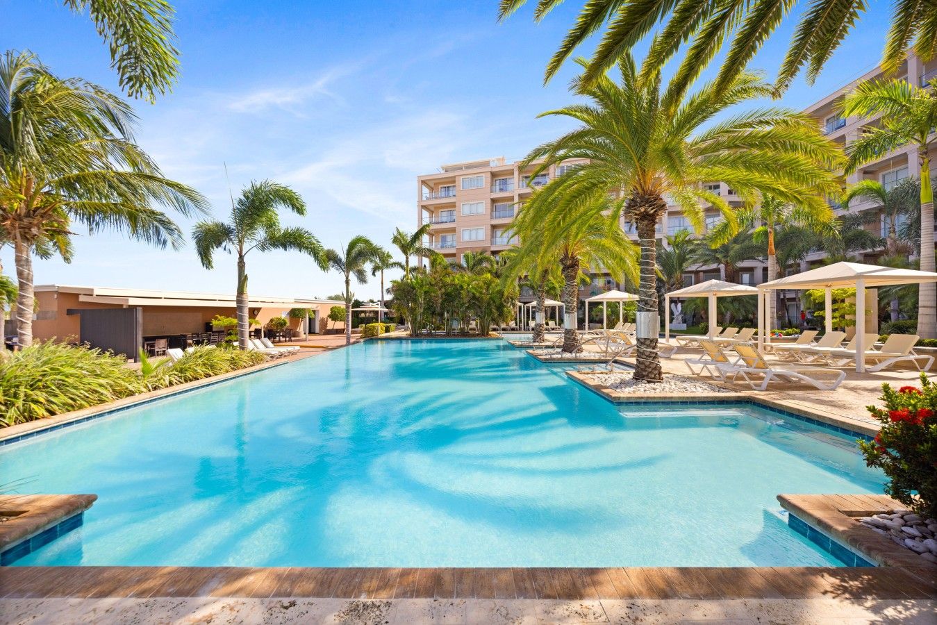 Piscina com espreguiçadeiras e palmeiras, cercada por um edifício com vários andares.