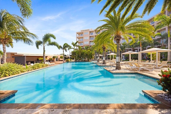 A swimming pool with lounge chairs and palm trees, surrounded by a building with multiple floors. - Luxe Beach Condo Le Vent Townhouse 17 by Bocobay (Noord)
