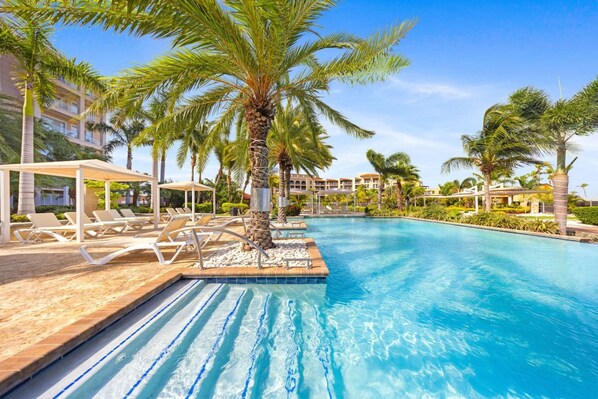 Une piscine entourée de chaises longues et de palmiers, le tout adossé à un bâtiment d’hôtel.