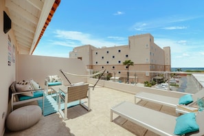 A balcony with a hammock, seating area, and a view of a building and the sea. - Luxe Beach Condo Le Vent Townhouse 17 by Bocobay (Noord)