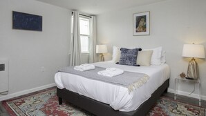 A neatly arranged bedroom with a large bed, two bedside lamps, a framed artwork above the bed, and a window with curtains. - The Cutler 1-bedroom · Near Mystic, Casinos + USCGA (Groton)