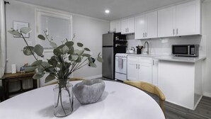 A modern kitchen with white cabinets, a stainless steel refrigerator, a microwave, and a sink. There is a vase with green leaves on the dining table. - The Cutler 1-bedroom · Near Mystic, Casinos + USCGA (Groton)