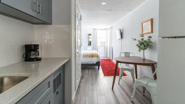 A compact kitchen with grey cabinets, a coffee maker, and a red rug leading to a dining area with a table and chairs. - The Kelly Studio · Near Mystic, Casinos + USCGA (Groton)