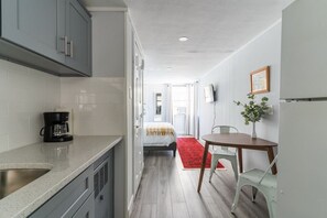 A compact kitchen with grey cabinets, a coffee maker, and a red rug leading to a dining area with a table and chairs. - The Kelly Studio · Near Mystic, Casinos + USCGA (Groton)