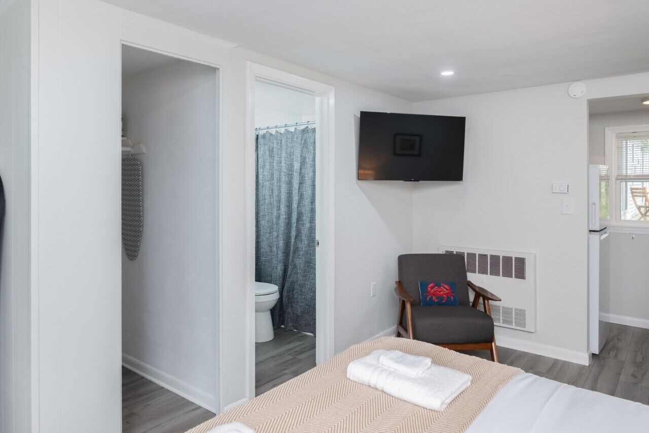 A modern hotel room with a bed, a chair, a TV, and a bathroom visible through an open door.