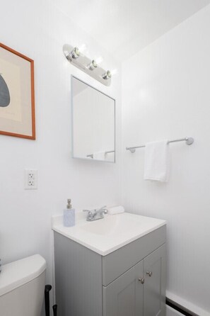 Un bany amb un lavabo blanc, un mirall amb il·luminació, una barra de tovalloles i una fotografia amb marc a la paret.
